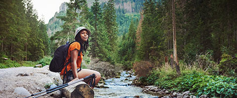 Person with backpack and hiking poles sitting on rock next to stream running through forest