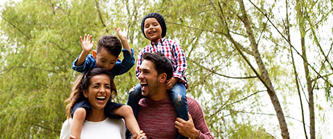 Cheerful adults laughing as two children sit on their shoulders with trees in the background.