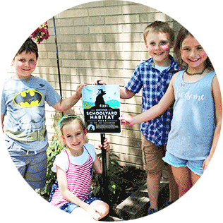 Four elementary school students standing and smiling together in the shade while holding a sign that reads Certified Schoolyard Habitat.